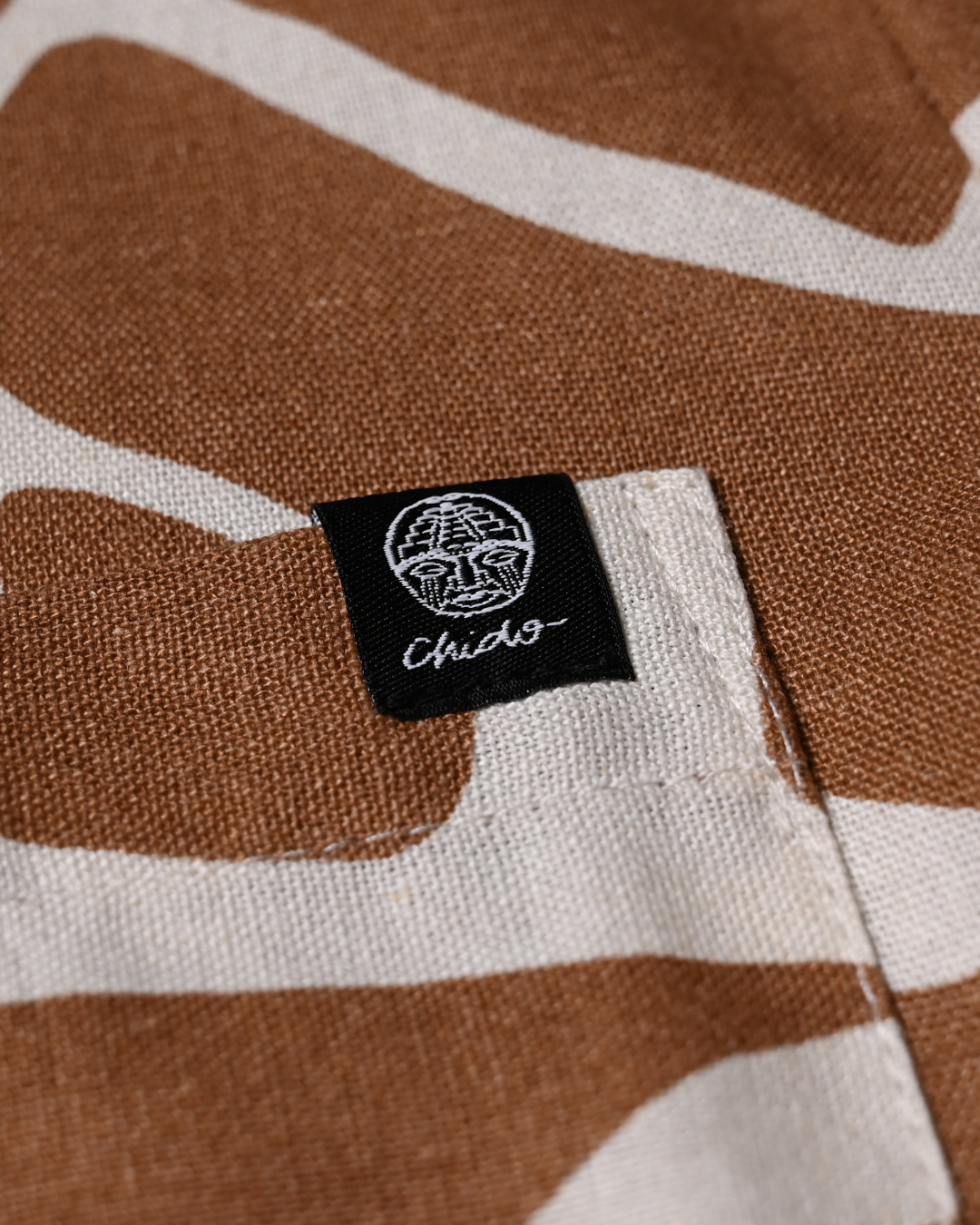 Detalle de tejido de lino y etiqueta CHIDO en camisa oversize beige con estampado exclusivo
